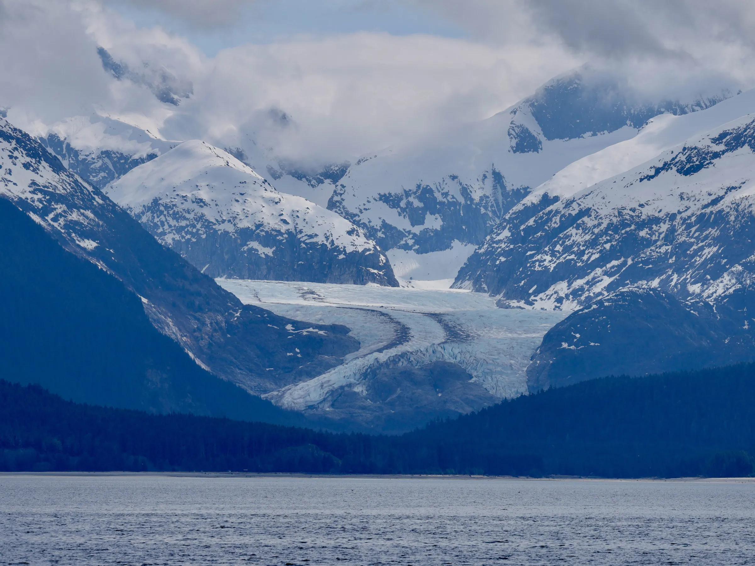 Snowy mountains with a glacier flowing between, under a cloudy sky.